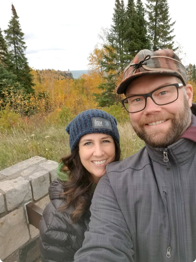 Close-up of Heather and her husband  Chad wearing fall gear and standing in front of a wooded area
